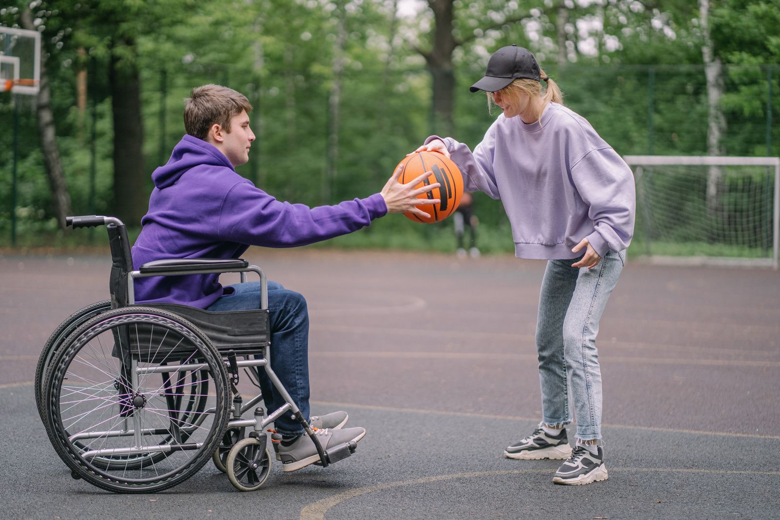A young man using a wheelchair passing a basketball to a friend on an outdoor court